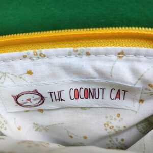 Pu&ograve; includere: Primo piano dell'interno di una pochette gialla con cerniera. La fodera &egrave; bianca con un motivo floreale. Un'etichetta con la scritta "THE COCONUT CAT" e un'illustrazione di un gatto &egrave; cucita all'interno della pochette. La cerniera &egrave; gialla e lo sfondo &egrave; verde.