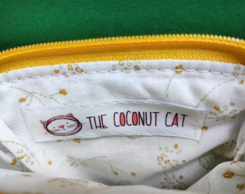 Pu&ograve; includere: Primo piano dell'interno di una pochette gialla con cerniera. La fodera &egrave; bianca con un motivo floreale. Un'etichetta con la scritta "THE COCONUT CAT" e un'illustrazione di un gatto &egrave; cucita all'interno della pochette. La cerniera &egrave; gialla e lo sfondo &egrave; verde.