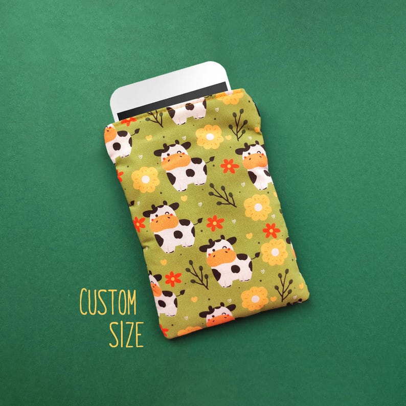 Pu&ograve; includere: Una custodia per telefono in tessuto verde con un motivo ripetuto di mucche dei cartoni animati, fiori arancioni e rametti. Il testo "CUSTOM SIZE" &egrave; stampato in giallo. Un telefono bianco &egrave; parzialmente visibile all'interno della custodia.