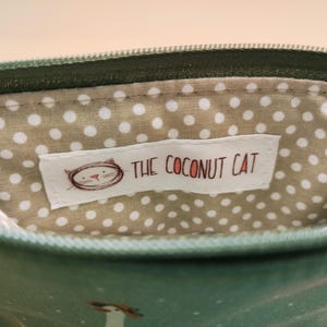 May include: Close-up of a small, open pouch with a green zipper and exterior. The interior features a beige fabric with white polka dots. A white label with the text "THE COCONUT CAT" and a cat illustration is visible inside the pouch.