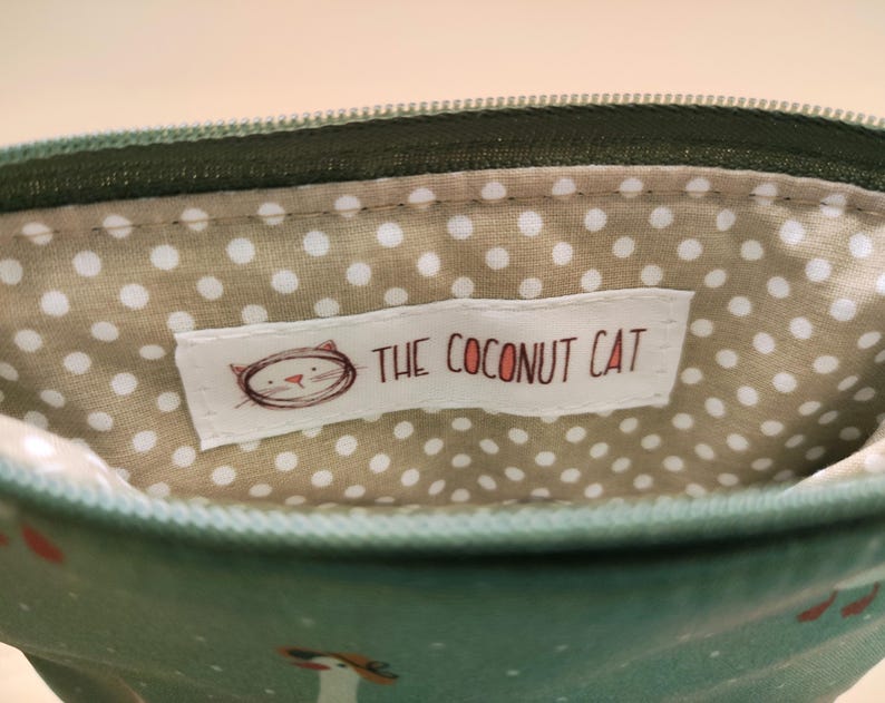 May include: Close-up of a small, open pouch with a green zipper and exterior. The interior features a beige fabric with white polka dots. A white label with the text "THE COCONUT CAT" and a cat illustration is visible inside the pouch.