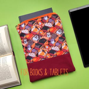 Könnte beinhalten: Eine rechteckige Buch- und Tablethülle mit einem bunten Muster von Katzen in Kisten. Die Hülle hat einen bordeauxfarbenen Boden und einen orangefarbenen Reißverschluss. Der Text "FOR BOOKS & TABLETS" ist unten aufgedruckt. Ein Buch und ein Tablet liegen daneben.