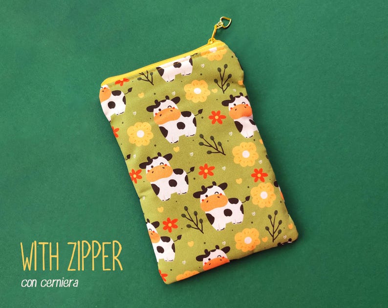 Pu&ograve; includere: Un astuccio rettangolare in tessuto verde con cerniera gialla e tiretto dorato. L'astuccio &egrave; decorato con mucche dei cartoni animati, fiori arancioni e ramoscelli. Il testo "WITH ZIPPER" &egrave; stampato in basso a sinistra.