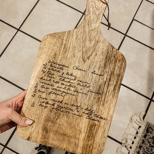 K&ouml;nnte beinhalten: Ein Holzbrett zum Schneiden mit einem handgeschriebenen Rezept f&uuml;r Wisconsin-K&auml;sesuppe. Das Rezept enth&auml;lt Zutaten wie Karotten, Sellerie, Zwiebeln, wei&szlig;e So&szlig;e, Margarine, Mehl, Milch und K&auml;se.