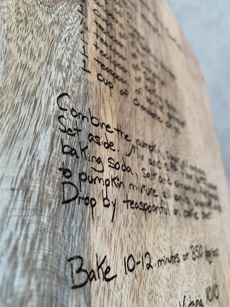 K&ouml;nnte beinhalten: Eine Nahaufnahme eines Holz-Schneidebretts mit einem handgeschriebenen Rezept f&uuml;r K&uuml;rbiskuchen. Das Rezept enth&auml;lt Zutaten wie K&uuml;rbis, Zucker, Salz, Backpulver und Vanilleextrakt. Die Anweisungen besagen, die Zutaten zu vermischen, beiseite zu stellen und 10-12 Minuten bei 350 Grad zu backen.