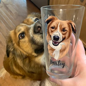 May include: A clear pint glass featuring a portrait of a dog with brown, white, and black fur. The dog's name, "Huxley," is written in black script at the bottom of the glass. A dog with similar coloring is in the background.