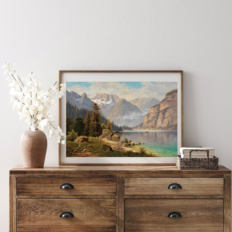 May include: A framed landscape painting depicting a mountain lake scene with a forest in the foreground and snow-capped mountains in the background.