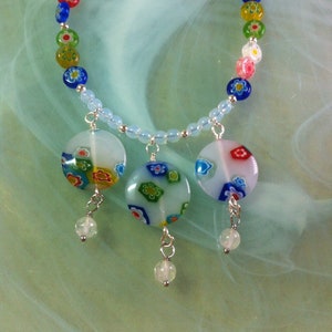 May include: A necklace with a light blue, white, and multi-colored glass bead design. The necklace has three large, round, white glass beads with multi-colored floral patterns hanging from it.
