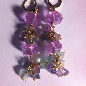 May include: A pair of dangle earrings with purple and blue gemstone beads. The earrings are made with gold wire and have a delicate, feminine look.
