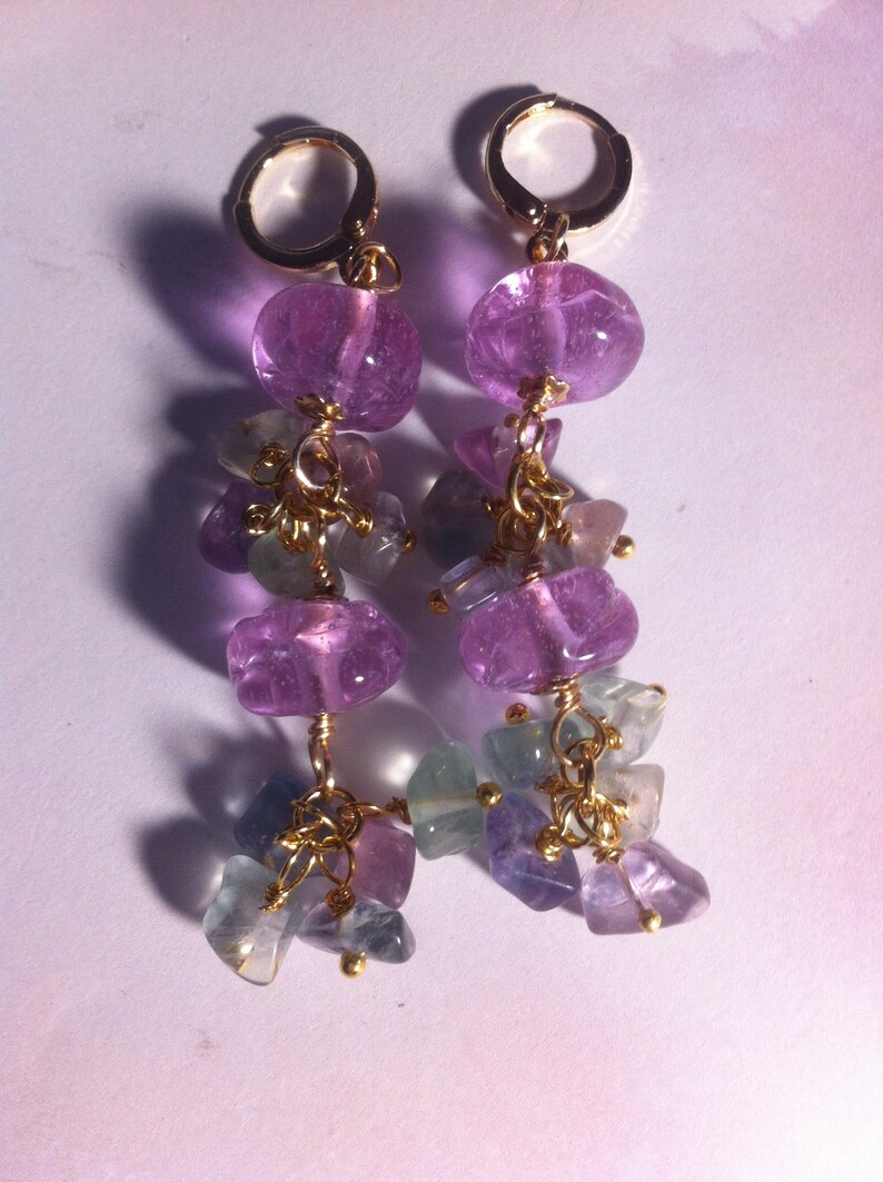 May include: A pair of dangle earrings with purple and blue gemstone beads. The earrings are made with gold wire and have a delicate, feminine look.