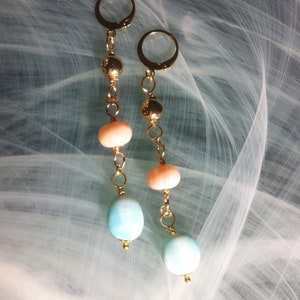Peut inclure: Paire de boucles d'oreilles pendantes avec des détails en métal doré. Chaque boucle d'oreille comprend un anneau doré, une chaîne avec des perles dorées, des perles couleur pêche et des perles ovales bleu clair. Les boucles d'oreilles sont présentées sur un fond blanc et bleu.