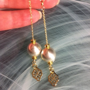 Puede incluir: Elegantes pendientes dorados con un diseño colgante. Cada pendiente presenta una gran cuenta de color plateado, una pequeña cuenta dorada y un dije dorado decorativo. Los pendientes están suspendidos de una delicada cadena dorada.