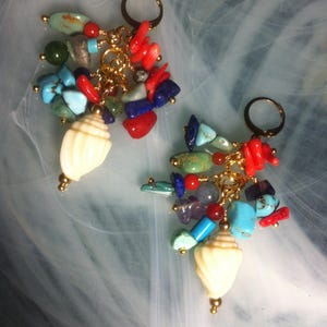 May include: A pair of dangle earrings with a collection of colourful gemstones and a cream seashell pendant. The earrings have gold-coloured hoops and chains, with red, blue, green, and turquoise stones.