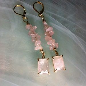 May include: A pair of gold earrings with pink rose quartz stones. The earrings are long and dangle, with a rectangular shaped rose quartz stone at the bottom.