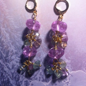 May include: A pair of dangle earrings with purple and green gemstone beads. The earrings are made with gold wire and have a gold hoop at the top.