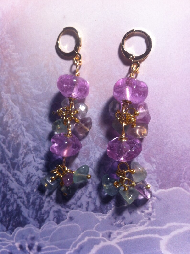 May include: A pair of dangle earrings with purple and green gemstone beads. The earrings are made with gold wire and have a gold hoop at the top.