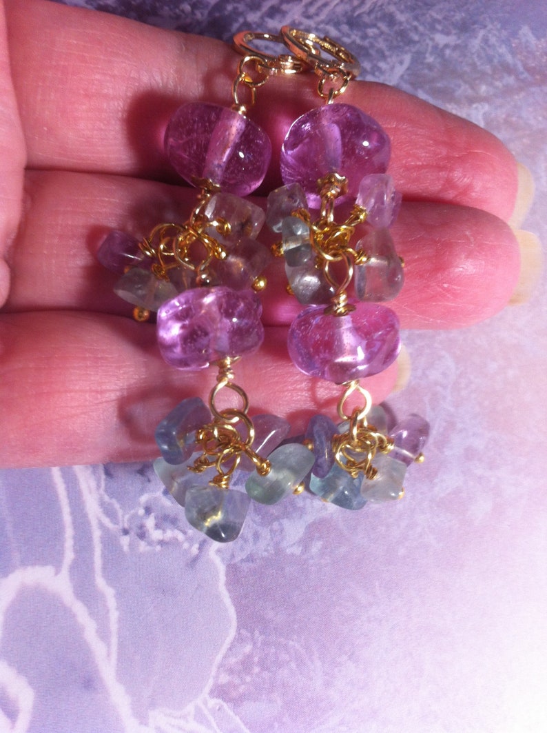 May include: A pair of dangle earrings with purple and green gemstone beads. The earrings are made with gold-toned metal and feature a cluster of smaller beads hanging from larger purple beads.