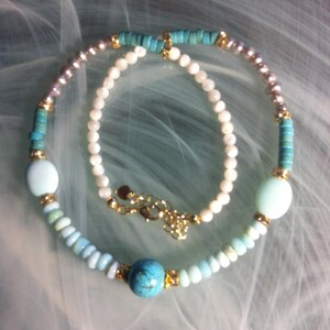 May include: A layered necklace featuring two strands. One strand consists of white beads with a gold-tone metal clasp. The second strand features turquoise-coloured disc beads, light blue round beads, a central turquoise stone, and metallic accents.
