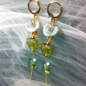Può includere: Un paio di orecchini pendenti dorati con perline di vetro verde e blu. Gli orecchini presentano un ciondolo a forma di fiore bianco con un centro blu.