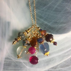 May include: A gold chain necklace featuring a cluster of colourful gemstone beads. The beads include clear, pink, blue, and brown stones, with gold accents. The necklace is set against a soft, blurred background.