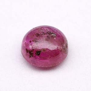 May include: A round, pink gemstone with a cabochon cut. The gemstone has a slightly cloudy appearance with some dark inclusions.