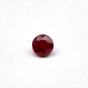 May include: A round, red gemstone with a faceted cut. The gemstone is set against a white background.