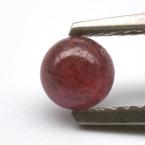 May include: A round, red gemstone held by tweezers. The gemstone is smooth and has a slight sparkle.