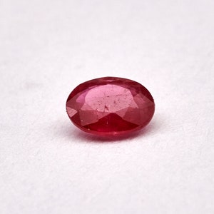 Puede incluir: Una piedra preciosa ovalada de color rojo oscuro con facetas. La piedra preciosa está sobre un fondo blanco.