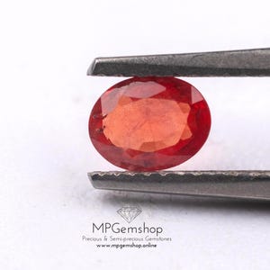 May include: A faceted, oval-shaped, red gemstone is held by tweezers. The gem has a warm, reddish-orange colour. The image includes the text "MPGemshop" and the website address. The gem is likely a precious stone.