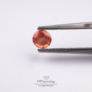 May include: A circular, orange gemstone is held by tweezers. The gemstone has a translucent quality with internal features. The background is plain white. The bottom of the image shows the text "MPGemshop Precious & Semi-precious Gemstones www.mpgems.shop.online".