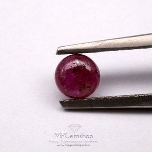 May include: A round, red gemstone with a slightly rough surface. The gemstone is held in tweezers.