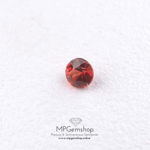 Op de afbeelding: Een ronde, gefacetteerde, dieprode edelsteen met een briljant slijpvorm, die interne lichtreflecties en helderheid vertoont. Onder de steen staat de tekst: MPGemshop Precious & Semi-precious Gemstones www.mpgemshop.online met een diamanticoon.