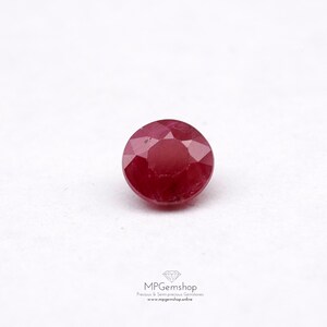 May include: A round, red gemstone with a faceted cut. The gemstone is set against a white background.