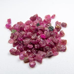 Può includere: Un mucchio di pietre preziose di rubino grezze, non tagliate, in varie tonalità di rosso e rosa. Le pietre grezze hanno una forma e una consistenza naturali e irregolari, con alcuni accenni di verde e marrone. Le pietre preziose sono sparse su una superficie bianca.