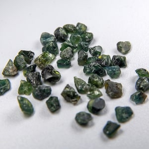 Puede incluir: Un conjunto de gemas en bruto, sin tallar, dispersas sobre una superficie blanca. Las piedras, de tamaños y formas variadas, exhiben tonos verdes y verde-azules. La textura rugosa y el aspecto natural de las gemas son notables.