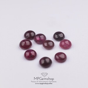 May include: Nine small, round gemstones in shades of red and purple are displayed on a white surface. The image features the text "MPGemshop Precious & Semi-precious Gemstones". The gemstones are likely cabochons.