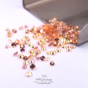 May include: A collection of small, round gemstones in shades of gold, orange, pink, and brown are scattered on a white surface. A metal scoop holds more of the gems. The image includes the text "MPGemshop Precious & Semi-precious Gemstones".