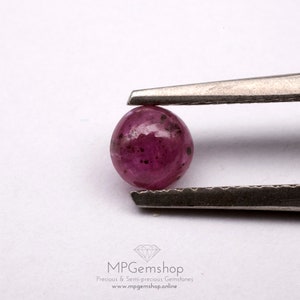 Peut inclure: Une pierre précieuse de rubis ronde, rose avec des inclusions foncées, tenue par des pinces sur un fond blanc.