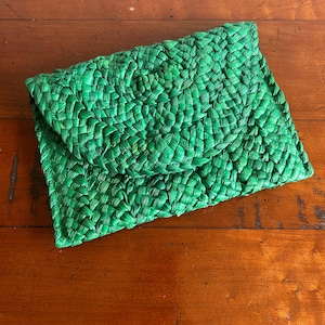 Könnte beinhalten: Eine hellgrüne gewebte Clutch-Tasche mit Klappe. Die Tasche besteht aus einem strukturierten Material, möglicherweise Bast oder einer ähnlichen Naturfaser. Die Clutch ist rechteckig mit einem abgerundeten Klappverschluss und liegt auf einer Holzoberfläche.