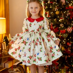 Puede incluir: Una niña pequeña con un vestido blanco con cuello rojo y estampado floral se para en una silla de madera frente a un árbol de Navidad decorado. Lleva una diadema con un diseño de asta de reno y sonríe a la cámara.