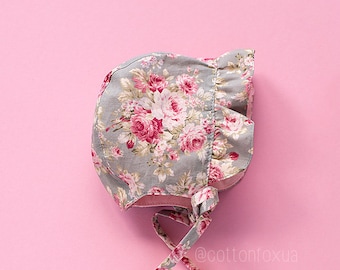 May include: A gray and pink floral bonnet with a ruffled edge and a tied bow. The bonnet has a floral pattern with pink roses and green leaves.