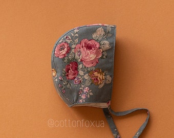 May include: A gray baby bonnet with a floral pattern featuring pink and red roses, yellow flowers, and green leaves. The bonnet has a tie closure and is set against an orange background. The text "@cottonfoxua" is visible.