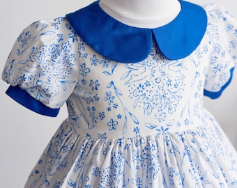 Könnte beinhalten: Ein weißes Kleid mit blauem Blumenmuster und einem blauen Peter-Pan-Kragen. Das Kleid hat Puffärmel und einen vollen Rock.