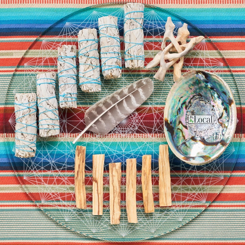 May include: A glass plate with a white geometric design on a red and blue striped background. The plate contains various items for smudging, including white sage bundles, palo santo sticks, a feather, and a seashell bowl with the word "Local" printed on it.