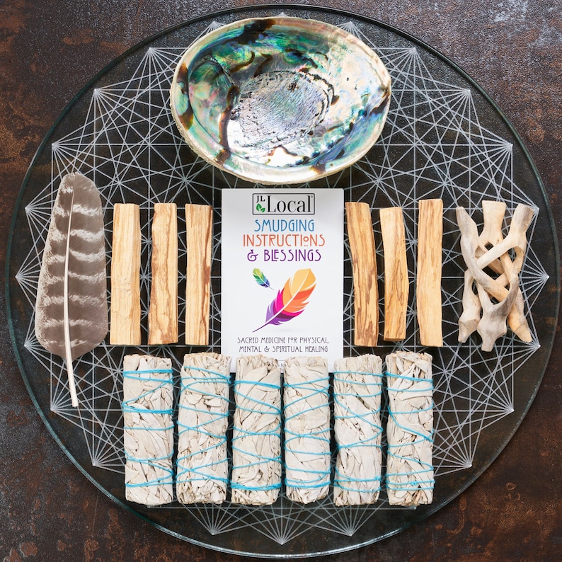 May include: A white sage smudging kit with four bundles of white sage, four Palo Santo sticks, a feather, a seashell, and a card with instructions and blessings. The kit is on a glass plate with a geometric design.