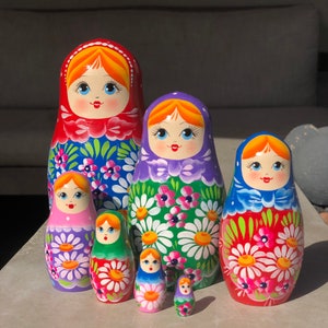 May include: A set of six traditional Russian nesting dolls, or matryoshka dolls. The dolls are painted in bright colors with floral patterns and feature smiling faces. The largest doll is red with blue and white flowers, the second largest is purple with green and white flowers, the third largest is red with green and white flowers, the fourth largest is pink with purple and white flowers, the fifth largest is green with red and white flowers, and the smallest doll is purple with white flowers.