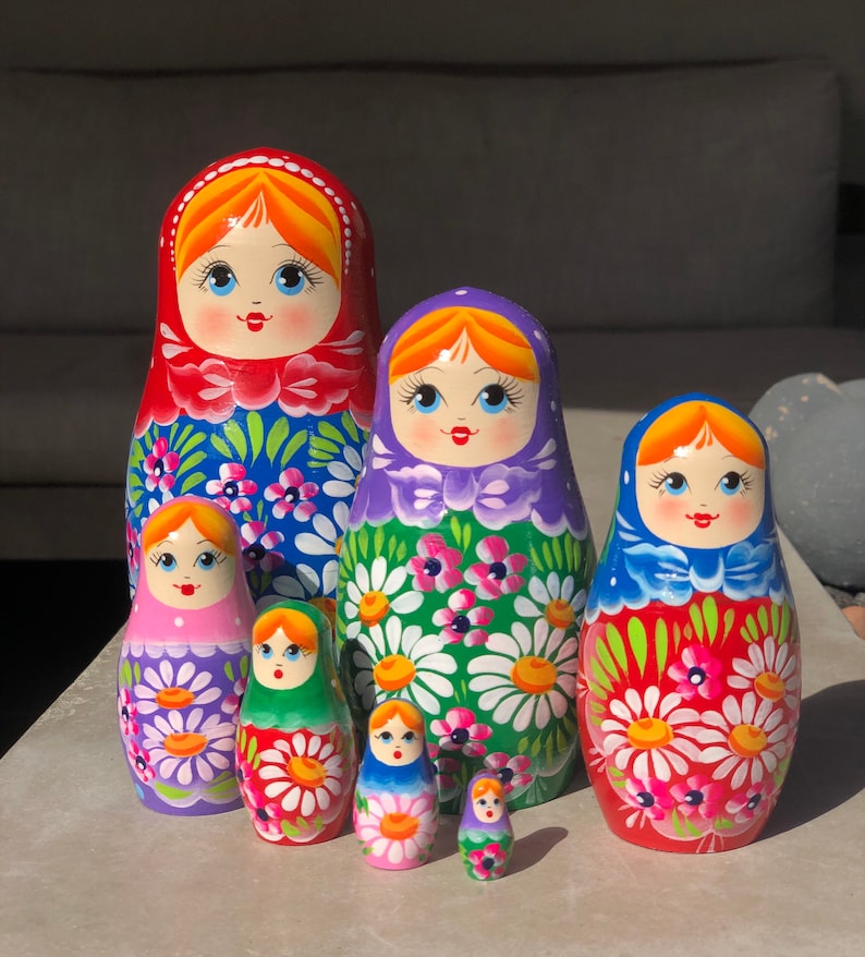 May include: A set of six traditional Russian nesting dolls, or matryoshka dolls. The dolls are painted in bright colors with floral patterns and feature smiling faces. The largest doll is red with blue and white flowers, the second largest is purple with green and white flowers, the third largest is red with green and white flowers, the fourth largest is pink with purple and white flowers, the fifth largest is green with red and white flowers, and the smallest doll is purple with white flowers.