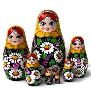 May include: A set of seven Russian nesting dolls, or Matryoshka dolls, of varying sizes. Each doll features a painted face, a yellow headscarf, and a black dress adorned with white daisies and purple accents. The dolls are arranged in a group.