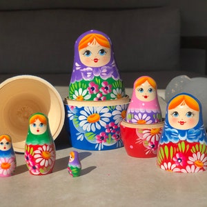 May include: A set of colorful Russian nesting dolls, also known as matryoshka dolls. The dolls are painted with floral patterns and feature smiling faces. The largest doll is blue with white flowers, and the smallest doll is purple with white flowers.