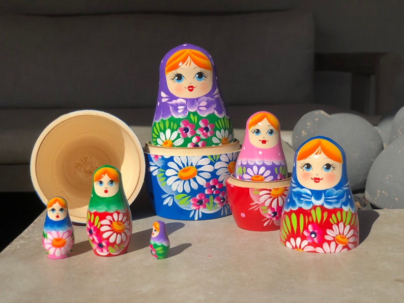 May include: A set of colorful Russian nesting dolls, also known as matryoshka dolls. The dolls are painted with floral patterns and feature smiling faces. The largest doll is blue with white flowers, and the smallest doll is purple with white flowers.